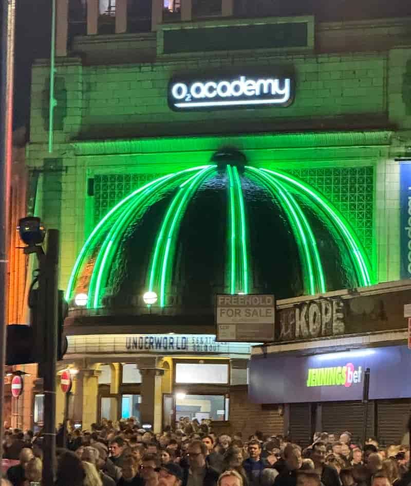 The dome at the entrance of Brixton Academy there are green lights of the dome and below it reads: Underworld 5, 6 & 7 February Sold Out. In front there are 1000s of smiling people leaving the venue